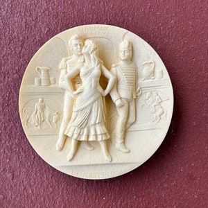 May include: A white ceramic plate with a raised relief sculpture of three figures from the opera Carmen. The figures are a man in a military uniform, a woman in a flowing dress, and another man in a suit. The plate is titled "Carmen" and "Bizet's "La" Davoliera" in raised lettering.