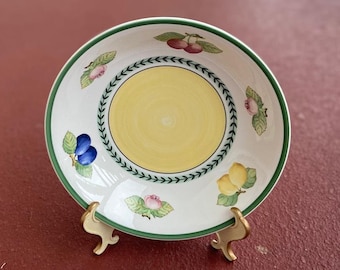 May include: A white ceramic bowl with a green rim and a yellow center. The bowl is decorated with a floral pattern and fruit, including lemons, cherries, and plums.