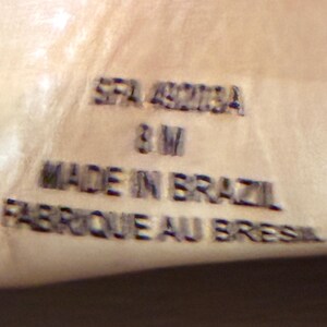 May include: A light tan leather surface featuring embossed black text that reads SFA 49218A, 8 M, MADE IN BRAZIL, and FABRIQUE AU BRESIL. The lettering is clearly stamped into the material, providing product information in a clear, legible font style.
