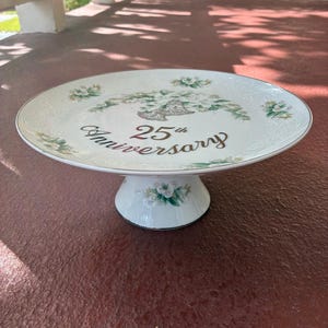 May include: A white ceramic cake stand with a floral design and the text "25th Anniversary" in gold script. The stand has a round top and a pedestal base, perfect for displaying a celebratory cake. The floral design includes green and white flowers.