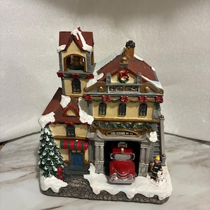May include: A miniature Christmas village fire station with a red fire truck in the garage. The building has a red roof, snow-covered accents, and a Christmas tree. The text "FIRE STATION NO. 24" is above the garage.