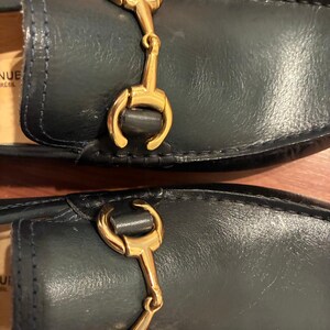 May include: A pair of dark navy blue leather loafers featuring a polished gold-tone metal horsebit detail across the vamp. The interior lining is tan, and the partial text visible on the inner sole reads NUE and BRASIL in black lettering.