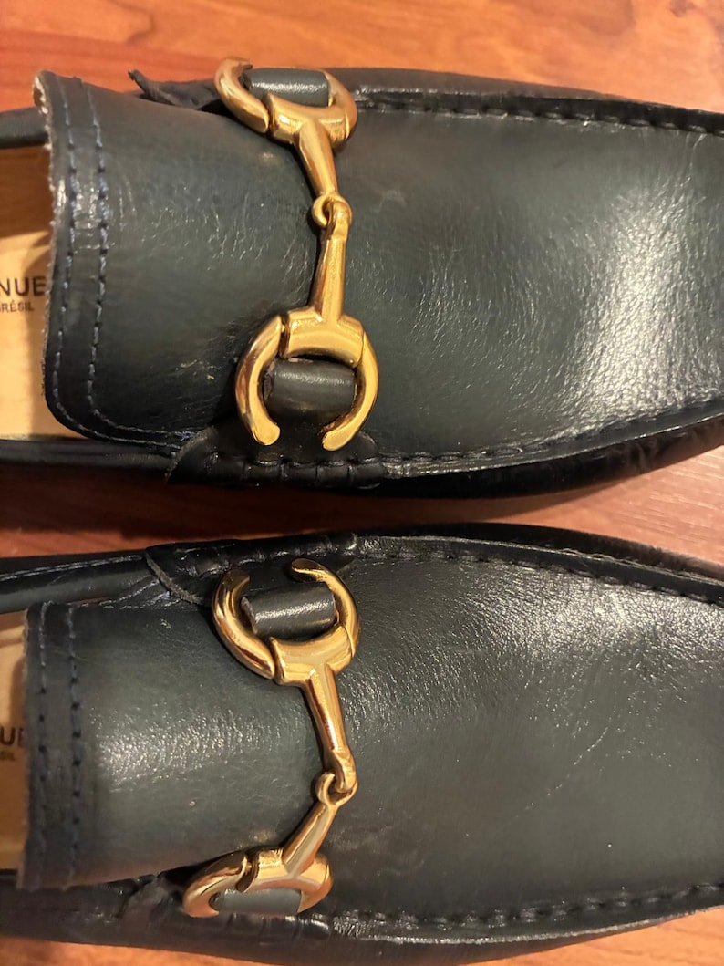 May include: A pair of dark navy blue leather loafers featuring a polished gold-tone metal horsebit detail across the vamp. The interior lining is tan, and the partial text visible on the inner sole reads NUE and BRASIL in black lettering.