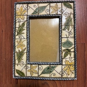 May include: Rectangular picture frame with a silver rope-like border. The frame's inner border is made of small, square tiles with leaf designs in green and yellow against a cream background. The frame is empty.