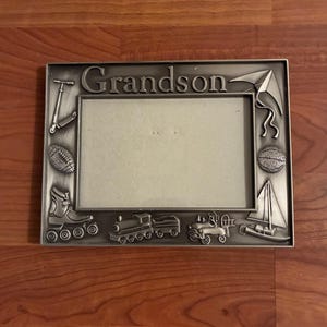 May include: Silver-colored metal picture frame with the word "Grandson" at the top. The frame has embossed images of a scooter, football, roller skate, train, car, sailboat, kite, and basketball. The frame is rectangular.