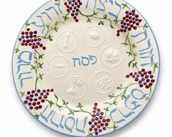 May include: A round ceramic Seder plate with a cream-colored base and a light blue rim. The border features painted purple and red grape clusters with green vines. Hebrew text is written in light blue, and six embossed circular icons represent traditional Passover foods, with the word Pesach in the center.
