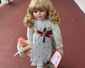 May include: A porcelain doll with blonde curly hair, wearing a white dress with a floral pattern and a red ribbon. The doll holds a basket of pink flowers and a card that says "Precious Moments".
