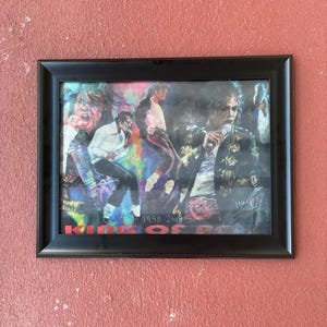 May include: A framed collage print featuring multiple depictions of Michael Jackson in various performance poses, with a colorful background, the text 1958-2009, and the words KING OF POP in bold red letters at the bottom of the rectangular black frame.