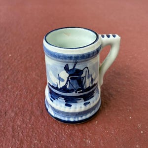 May include: Small, cylindrical ceramic mug with a handle. The mug features a blue and white windmill design, with a blue rim and base. The handle has a series of small, decorative holes. The mug is approximately 3 inches tall.