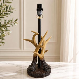 May include: A table lamp base featuring a central black metal rod and a socket, supported by a sculpted stand designed to resemble a cluster of light brown and dark brown deer antlers rising from a circular, textured, dark brown base.