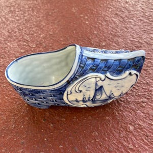 May include: Blue and white ceramic clog-shaped planter. The planter features a woven basket design on the sides and a nautical scene on the front. The interior is white.