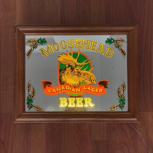 May include: A rectangular wooden-framed mirror featuring the Moosehead Canadian Lager Beer logo. The logo includes a gold moose head, the words "MOOSEHEAD", "CANADIAN LAGER", and "BEER" in yellow and orange.