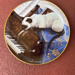 May include: Decorative plate featuring a Siamese cat resting on a blue cushion with a piano and sheet music. The plate has a gold rim and a signature at the bottom. The cat is white with dark brown markings.