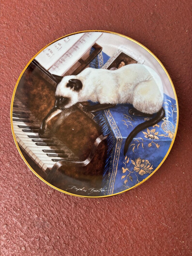 May include: Decorative plate featuring a Siamese cat resting on a blue cushion with a piano and sheet music. The plate has a gold rim and a signature at the bottom. The cat is white with dark brown markings.
