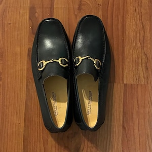 May include: A pair of black leather loafers featuring a gold-toned metal horsebit detail across the vamp. The interior soles are light tan and clearly printed with the text Saks Fifth Avenue, Made in Brazil, Fabrique au Bresil.