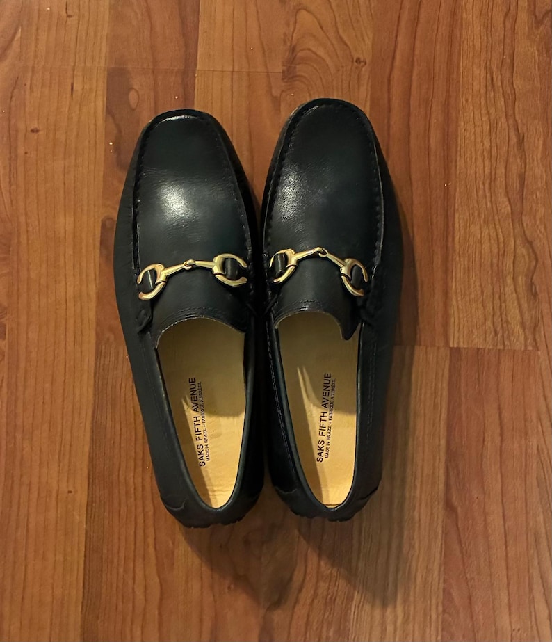 May include: A pair of black leather loafers featuring a gold-toned metal horsebit detail across the vamp. The interior soles are light tan and clearly printed with the text Saks Fifth Avenue, Made in Brazil, Fabrique au Bresil.