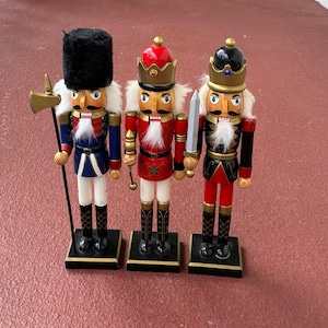 May include: Three decorative wooden nutcrackers in a row. Each nutcracker is painted with a different uniform design. The nutcrackers are wearing black boots and are standing on black bases. One holds an axe, another a sword, and the third a scepter.