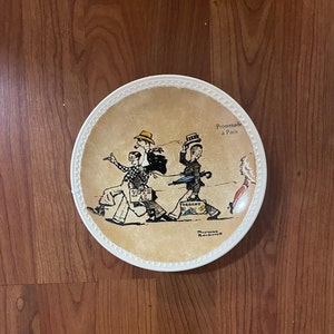 May include: A white ceramic plate with a brown background featuring a cartoon illustration of four people walking down a street in Paris. The text "Promenade a Paris" is printed on the plate. The illustration includes a person holding a sign that says "Vote for All".