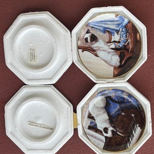 May include: Four decorative plates featuring Siamese cats in various settings. Each plate is displayed in a white, octagonal protective case. The plates have gold rims and detailed artwork, showcasing the cats with furniture and accessories.