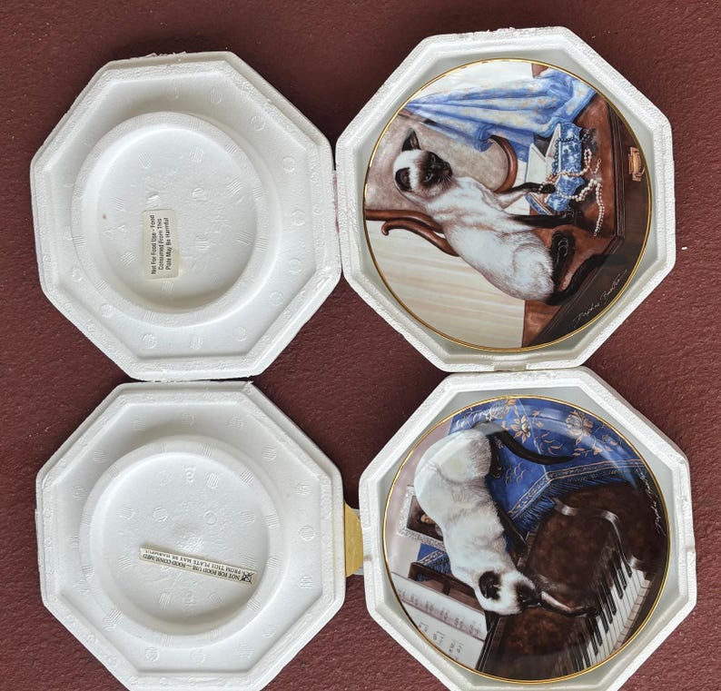 May include: Four decorative plates featuring Siamese cats in various settings. Each plate is displayed in a white, octagonal protective case. The plates have gold rims and detailed artwork, showcasing the cats with furniture and accessories.