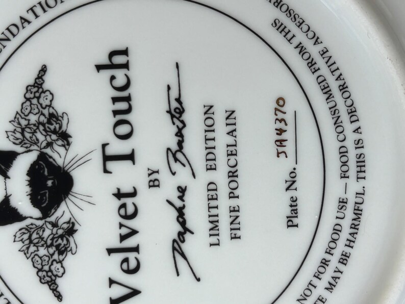 May include: White porcelain plate with a black and white Siamese cat illustration and floral accents. The plate is labeled "Velvet Touch" by Daphne Baxter, a limited edition fine porcelain piece. Plate number JA4370.