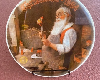 May include: A white ceramic plate with a Christmas 1984 design featuring a painting of Santa Claus working on a toy. The plate has a black metal stand and the signature "Norman Rockwell" at the bottom.