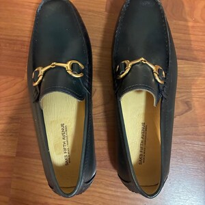 May include: A pair of dark navy blue leather loafers featuring a gold-tone metal horsebit detail across the vamp. The interior lining is light beige and features the text SAKS FIFTH AVENUE MADE IN BRAZIL - FABRIQUE AU BRESIL printed in black.