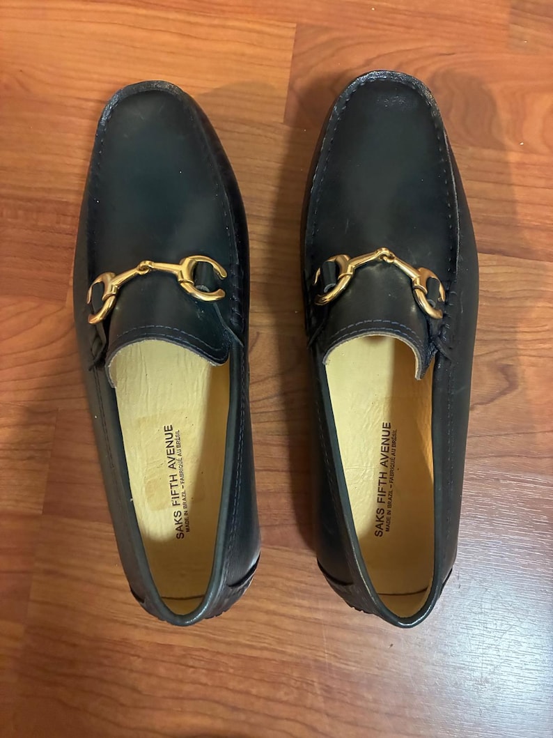 May include: A pair of dark navy blue leather loafers featuring a gold-tone metal horsebit detail across the vamp. The interior lining is light beige and features the text SAKS FIFTH AVENUE MADE IN BRAZIL - FABRIQUE AU BRESIL printed in black.