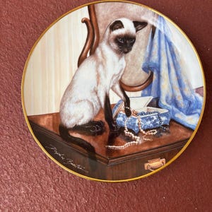 May include: Decorative plate featuring a Siamese cat sitting on a wooden surface. The cat is cream-colored with dark brown markings. A blue and white jewelry box and a pearl necklace are also on the surface. The plate has a gold rim and a signature.