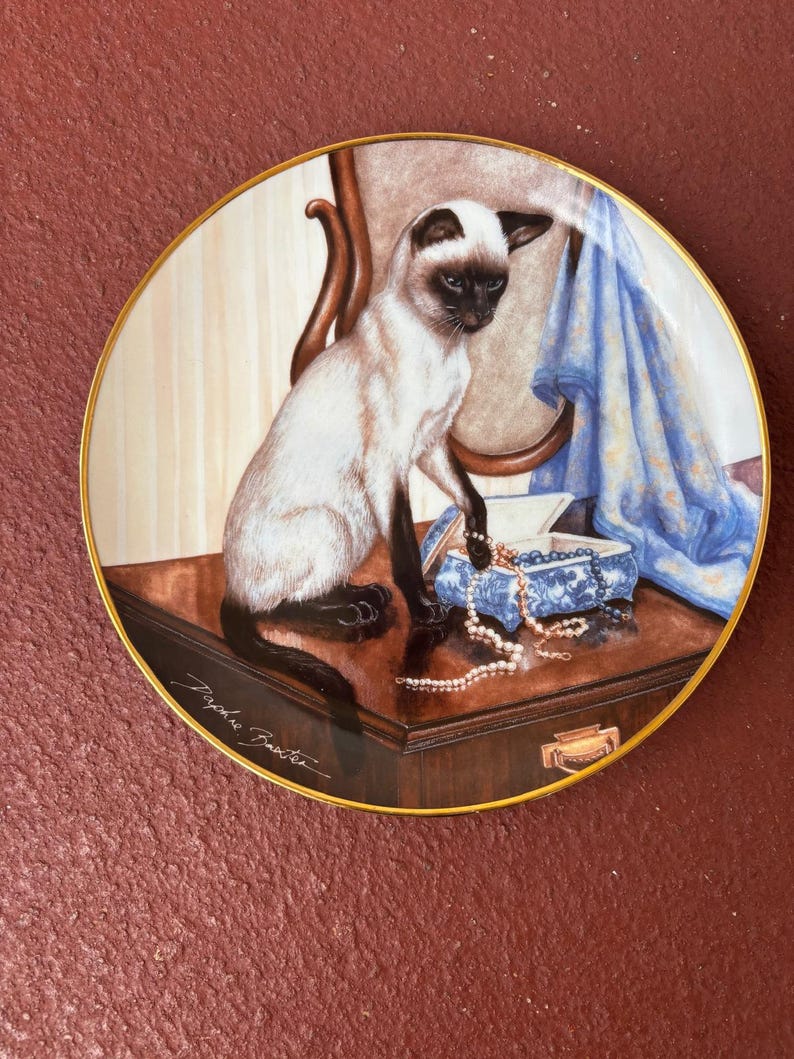May include: Decorative plate featuring a Siamese cat sitting on a wooden surface. The cat is cream-colored with dark brown markings. A blue and white jewelry box and a pearl necklace are also on the surface. The plate has a gold rim and a signature.