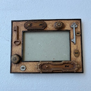 May include: A rectangular photo frame with a light brown wood-grain texture. The frame is decorated with various antique-style elements, including keys, keyholes, and decorative knobs. The frame's border is dark brown.