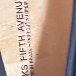 May include: A tan-colored shoe insole featuring the embossed text SAKS FIFTH AVENUE and MADE IN BRAZIL FABRIQUE AU BRESIL. The surface is smooth and light brown, showing the interior branding of the footwear with clear, legible lettering.