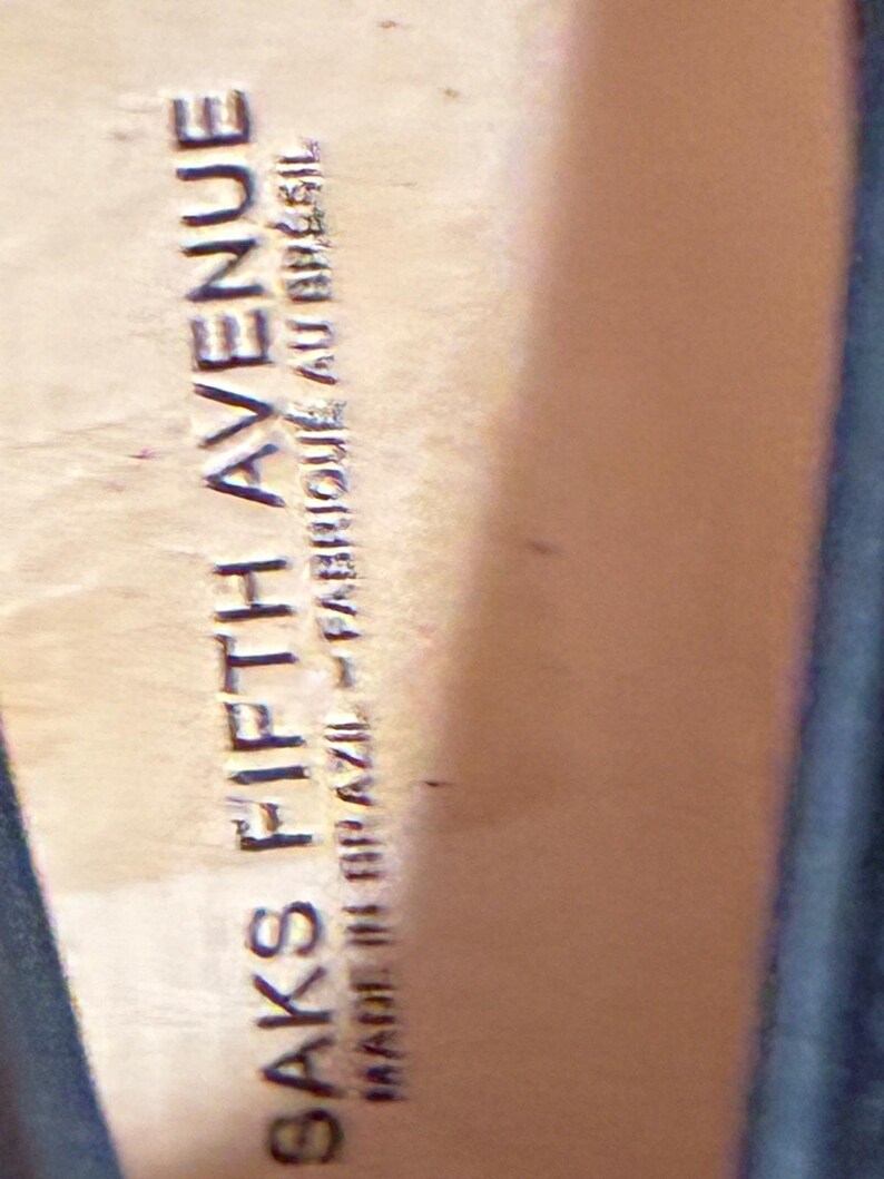 May include: A tan-colored shoe insole featuring the embossed text SAKS FIFTH AVENUE and MADE IN BRAZIL FABRIQUE AU BRESIL. The surface is smooth and light brown, showing the interior branding of the footwear with clear, legible lettering.