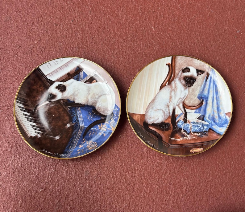 May include: Two decorative plates with gold rims. One plate depicts a Siamese cat on a piano, the other shows a cat on a table with a blue cloth and jewelry. The plates have a detailed, painted design.