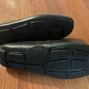 May include: A pair of black leather loafers featuring a distinct rubber sole design with segmented rectangular traction pads. The shoes are positioned to show the underside, highlighting the black rubber grip pattern and the smooth leather upper construction.
