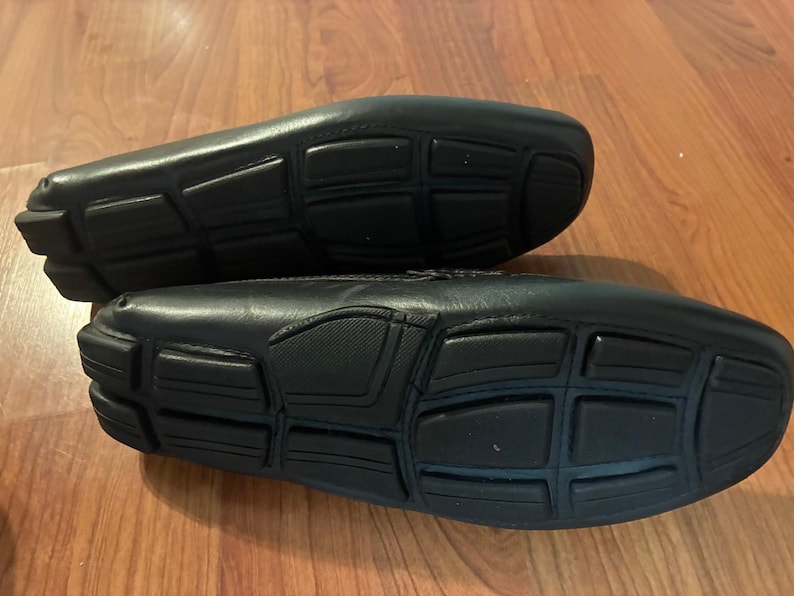 May include: A pair of black leather loafers featuring a distinct rubber sole design with segmented rectangular traction pads. The shoes are positioned to show the underside, highlighting the black rubber grip pattern and the smooth leather upper construction.