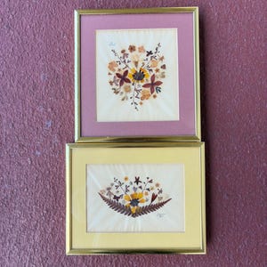 May include: Two framed floral art pieces. The top frame has a pink mat and the bottom frame has a yellow mat. Both frames have gold-colored metal frames. The floral arrangements feature dried flowers in shades of yellow, orange, and brown.