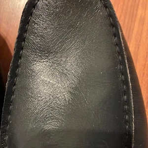May include: A black leather loafer shoe featuring a rounded toe and visible stitching along the perimeter of the vamp. The smooth, dark leather surface shows a subtle grain texture and a slight sheen, highlighting the construction of the footwear.