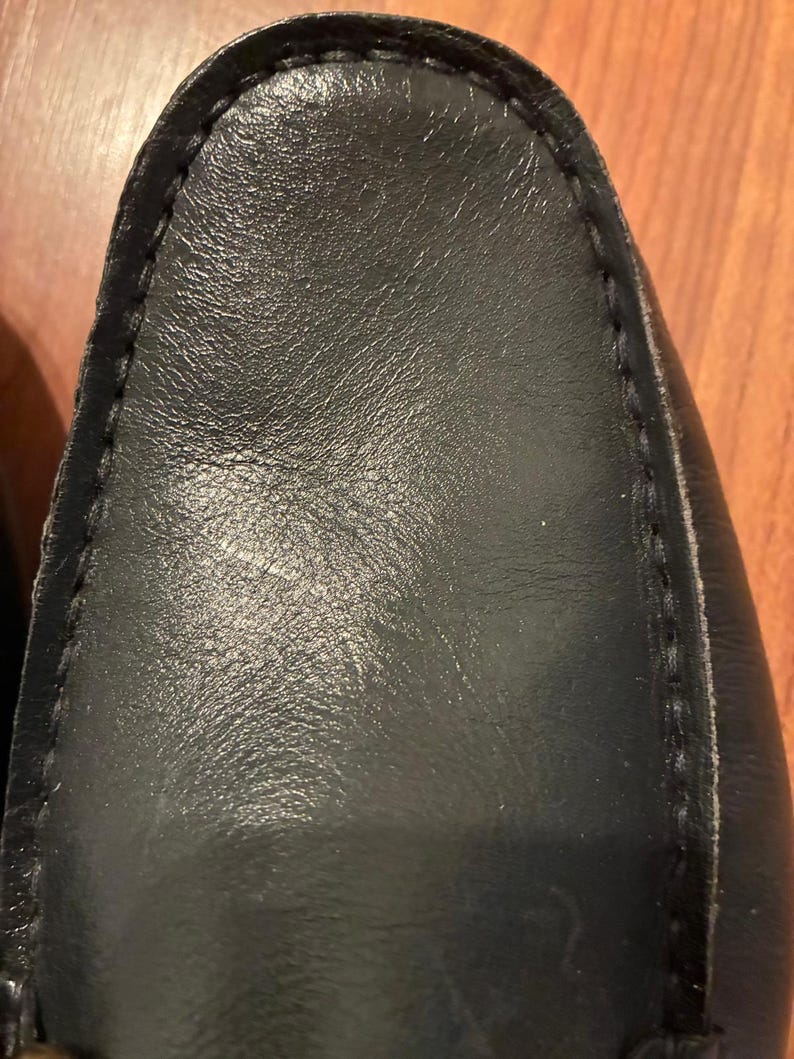 May include: A black leather loafer shoe featuring a rounded toe and visible stitching along the perimeter of the vamp. The smooth, dark leather surface shows a subtle grain texture and a slight sheen, highlighting the construction of the footwear.