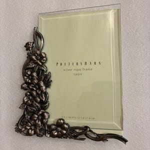 May include: A silver rope frame from Pottery Barn. The frame is rectangular with a clear glass front. The left side and bottom feature an ornate floral design. The frame is 5 x 7 inches (12.7 x 17.8 cm).