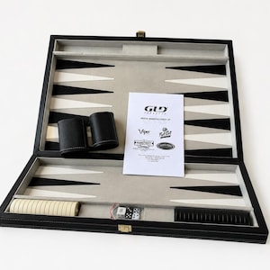 May include: Open backgammon set with black and white playing surface, black leather dice cups, dice, and ivory-colored game pieces. The set includes a printed sheet with logos. The case is black with a gold-colored clasp.