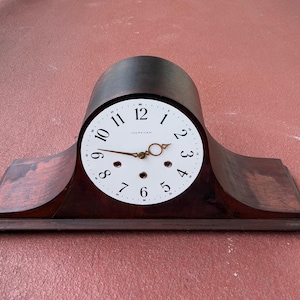 May include: A dark brown wooden mantel clock with a white clock face. The clock face has black numerals and gold-colored hands. The clock is rectangular with a curved top and is set against a red background.