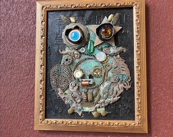 Może przedstawiać: Oprawione dzieło sztuki przedstawiające sowę w stylu steampunk. Sowa zbudowana jest z różnych metalowych przedmiotów, w tym kół zębatych, części zegarków i biżuterii. Oczy sowy są niebieskie i bursztynowe, a rama jest złota.