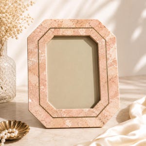 May include: A pink marble picture frame with an octagonal shape. The frame has a double border design and a neutral-colored interior. The frame is displayed on a marble surface.