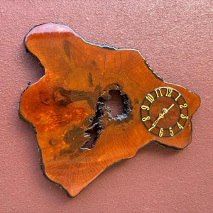 May include: A unique wooden wall clock with a natural, organic shape. The clock face is round with gold-colored numerals and hands. The wood has a rich, reddish-brown color with a glossy finish.