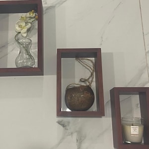 May include: Three wooden picture frames with different contents. The left frame has a white flower in a clear glass vase. The middle frame has a brown coconut shell hanging from a string. The right frame has a white candle with the text "Nature's Wick" on the label.
