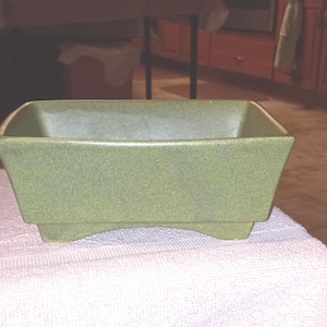 May include: A rectangular, green ceramic planter with a slightly textured surface. The planter has a flared design and sits on four small feet. The interior is a lighter shade of green.