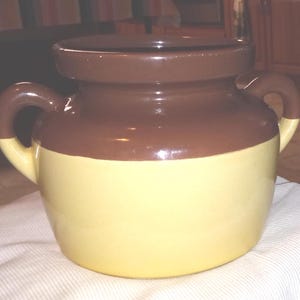 May include: A two-toned ceramic jar with two curved handles. The top and handles are brown, while the lower portion is yellow. The jar has a wide opening and a rounded shape, suitable for kitchen or decorative use.