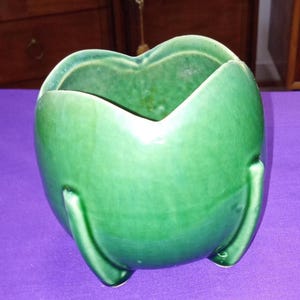May include: A green ceramic planter with a heart-shaped opening. The planter has three curved legs and a glossy finish. The interior is a darker shade of green. The planter is likely for indoor use.