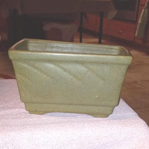 以下が含まれることがあります： 長方形の、薄緑色のセラミックプランター。側面にテクスチャ加工された斜め模様があります。プランターは少し斑点模様で、小さな足の上に置かれています。屋内または屋外の植物に最適です。